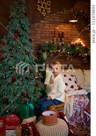 A young boy in a white sweater sits next to a Christmas tree and reaches out to carefully hang a red 131838695