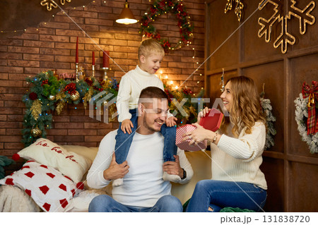 Happy family in white sweaters exchanging a Christmas gift in a cozy loft-style room with a brick Happy family in white sweaters exchanging a Christmas gift in a cozy loft-style room with a brick 131838720