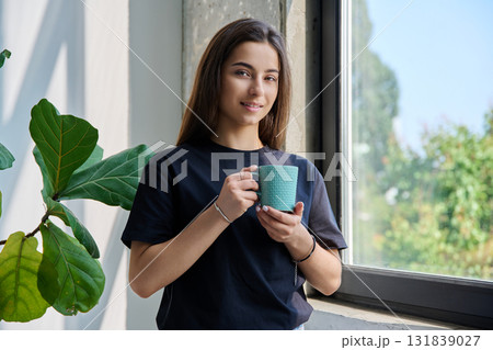 Portrait of smiling relaxed teenage girl posing with cup near window 131839027