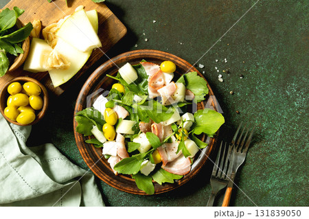 Fresh arugula salad with prosciutto, melon, and olives for a delicious summer meal. Top view flat lay. Fresh arugula salad with prosciutto, melon, and olives for a delicious summer meal. Top view flat lay. 131839050