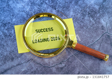A magnifying glass rests upon a yellow note that displays the message Success Loading 2026. The concept visualizes planning and strategizing for business success in the future 131841757