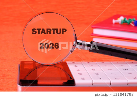 A calculator and books are positioned on a desk as a magnifying glass examines the concept of Startup 2026, implying research and financial planning for a business 131841798