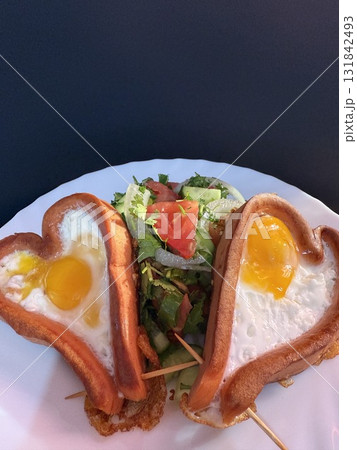 A close-up of two Frankfurt sausages, cut into hearts and fried with eggs in vegetable oil, and a vegetable salad. Cooking delicious food at home. 131842493
