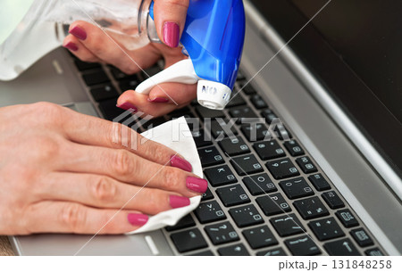 Woman cleaning laptop keyboard with white tissue, detail on her fingers holding paper towel, blue alcohol sprayer near - disinfection concept 131848258