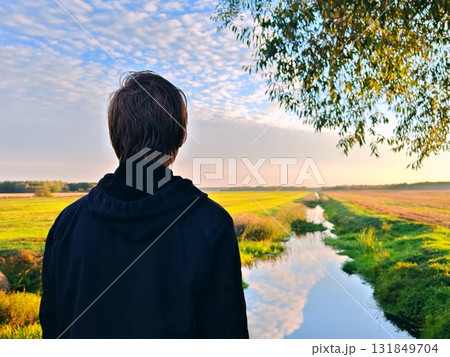 Guy is standing with his back to camera in an autumn landscape in countryside. A man in a hoodie stands with his back to the camera against a beautiful forest lake. Enjoy nature and travel Guy is standing with his back to camera in an autumn landscape in countryside. A man in a hoodie stands with his back to the camera against a beautiful forest lake. Enjoy nature and travel 131849704
