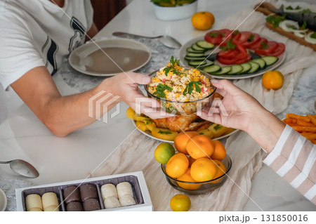 Close-up of hands holding a bowl of Olivier salad above a festive holiday table with roasted chicken, mandarins, and sweets. Cozy Christmas and New Year celebration with traditional dishes and warm 131850016