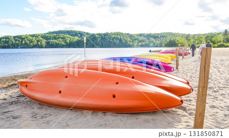 Colorful kayaks lined up on sandy beach by tranquil lake, surrounded by lush greenery and soft clouds, inviting outdoor adventure and relaxation 131850871