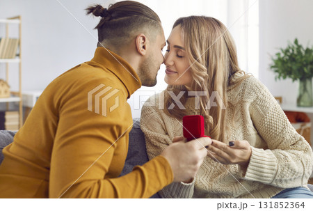 Young man with a red gift box proposing to his happy girlfriend sitting on the sofa. Young man with a red gift box proposing to his happy girlfriend sitting on the sofa. 131852364
