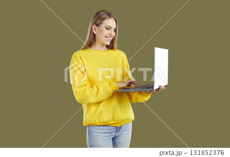 Smiling blondy woman in casual yellow sweatshirt and jeans working on laptop on khaki background. Smiling blondy woman in casual yellow sweatshirt and jeans working on laptop on khaki background. 131852376