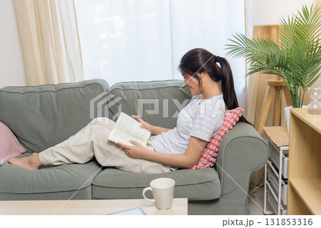 Smiling young woman teen person reading book on comfortable sofa, resting in cozy room, enjoying peaceful moment at home with refreshing beverage near green plant. Smiling young woman teen person reading book on comfortable sofa, resting in cozy room, enjoying peaceful moment at home with refreshing beverage near green plant. 131853316