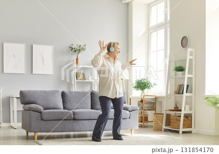 Cheerful mature woman with blonde hair wearing headphones dancing in bright modern living room. 131854170