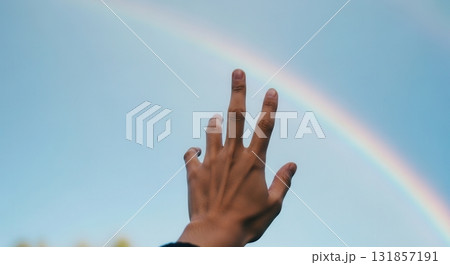 Human hand reaching out towards a vibrant rainbow arching across a clear blue sky 131857191