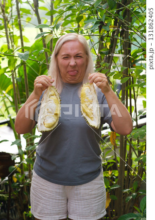 Woman holding durian fruit in a lush tropical garden while making a funny face due to its strong smell Woman holding durian fruit in a lush tropical garden while making a funny face due to its strong smell 131862405