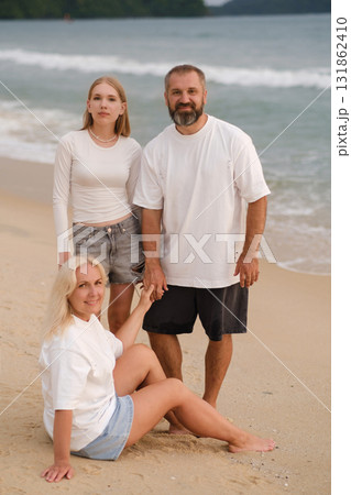 Family enjoys a sunny day at the beach, smiling while standing together by the shoreline with gentle waves in the background 131862410
