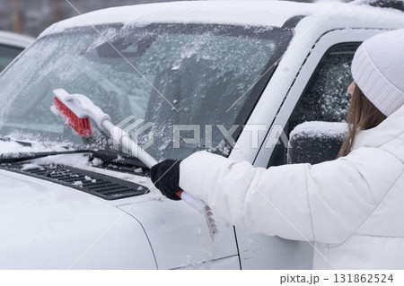 Removing the accumulation of snow from a vehicle on a chilly winter day requires effort , A woman in a white jacket cleans snow and ice with a brush from the windshield of a white car in winter, a 131862524