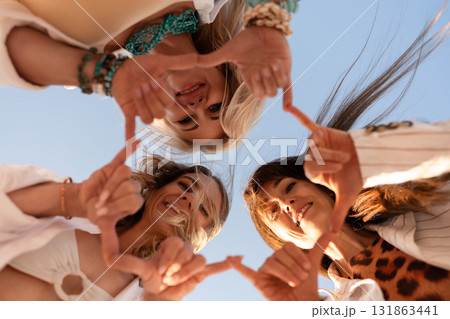 Women, friendship, happiness. Three joyful friends forming heart shapes with their hands against a clear blue sky, celebrating togetherness and love. 131863441