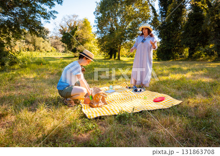 Middle-aged mother and her young Caucasian daughter relaxing on a blanket in a summer park enjoying healthy snacks and family recreation outdoors. Wide shot 131863708