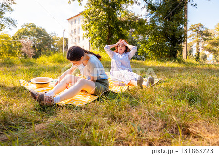Family picnic photo with visible bug irritation on woman and daughter relaxing on soft blanket in grassy park during hot season. Low angle view Family picnic photo with visible bug irritation on woman and daughter relaxing on soft blanket in grassy park during hot season. Low angle view 131863723