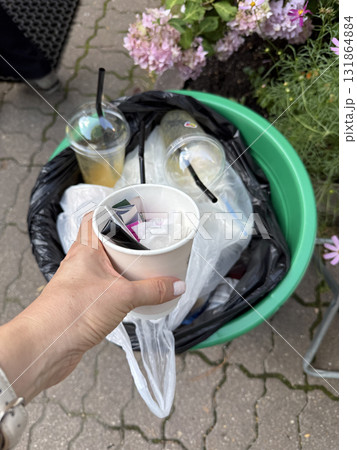 Hand disposing of a paper cup into green recycling bin on a street with flowers in background Hand disposing of a paper cup into green recycling bin on a street with flowers in background 131864884