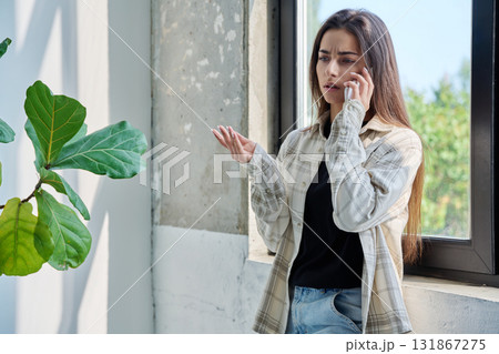 Serious teenage female standing at home talking on smartphone Serious teenage female standing at home talking on smartphone 131867275