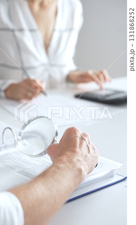 Close up of accountant hand using a magnifying glass and a calculators, to audit financial reports in brightly lit office. Audit and taxes concept 131868252