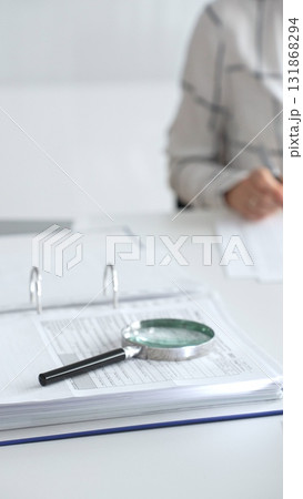 Magnifying glass on financial documents is in focus while an accountant using a calculator and taking notes, emphasizing accounting, finance, close up vertical view. Audit and taxes 131868294