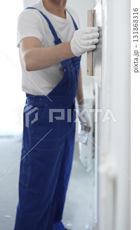 Man construction worker wearing protective gloves and blue construction coveralls, spreading plaster smoothly across wall using professional drywall taping knife Man construction worker wearing protective gloves and blue construction coveralls, spreading plaster smoothly across wall using professional drywall taping knife 131868316