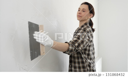 Woman construction professional wearing checkered shirt is smoothing white wall surface, applying putty with professional spatula technique, showcasing skilled interior renovation work Woman construction professional wearing checkered shirt is smoothing white wall surface, applying putty with professional spatula technique, showcasing skilled interior renovation work 131868432