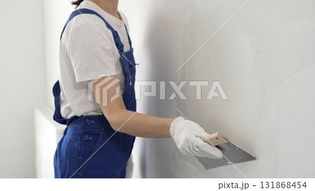 Unknown construction worker woman in blue overalls smoothing wall surface, applying plaster with putty knife, wearing protective white gloves during home renovation work Unknown construction worker woman in blue overalls smoothing wall surface, applying plaster with putty knife, wearing protective white gloves during home renovation work 131868454