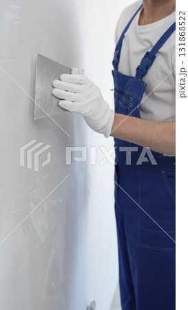 Close-up of man construction worker wearing gloves and blue overalls is using a putty knife to apply plaster on a white wall, performing home renovation and improvement 131868522