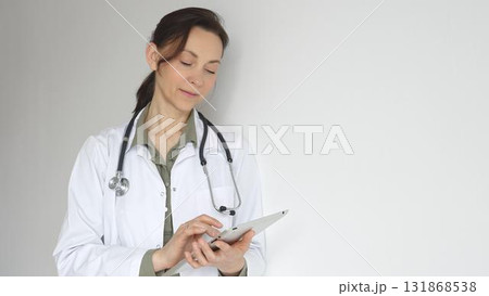 Doctor woman wearing white medical coat over green blouse, stethoscope, is reviewing patient records on digital tablet, standing in medical workspace with natural lighting. Medicine concept Doctor woman wearing white medical coat over green blouse, stethoscope, is reviewing patient records on digital tablet, standing in medical workspace with natural lighting. Medicine concept 131868538