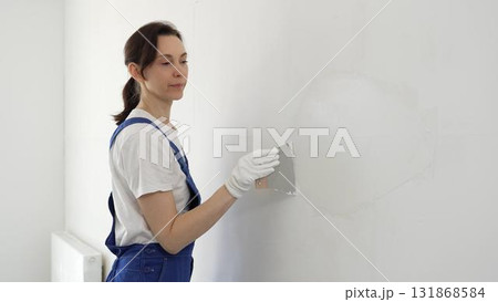 Female construction worker spreading finishing putty on white wall, wearing protective gloves and blue work overall while smoothing surface with putty knife for precise home repair 131868584
