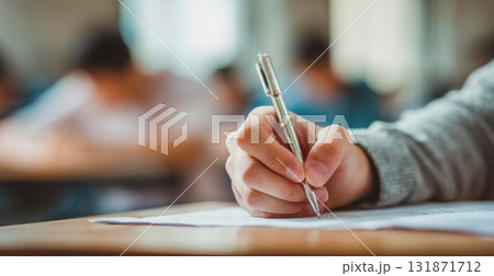 Close-up of a hand holding a pen and paper, doing an exam in school, writing something. Blurred background. The student is sitting at their desk, taking a mental test for a final-year class. 131871712
