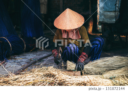Vietnamese woman in traditional straw hat cuts reeds at a woven mat factory in a village in Vietnam Vietnamese woman in traditional straw hat cuts reeds at a woven mat factory in a village in Vietnam 131872144