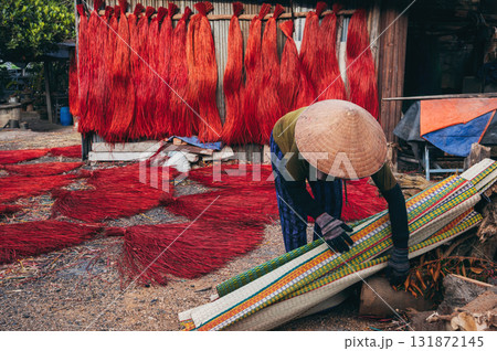 Vietnamese woman in traditional straw hat works on background of dyed reeds for production of woven mats in a factory in an artisan village in Vietnam Vietnamese woman in traditional straw hat works on background of dyed reeds for production of woven mats in a factory in an artisan village in Vietnam 131872145