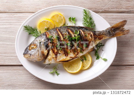 Grilled fish on a white plate, top view, wooden backdrop. Fried fish with slice of lemon and fresh herbs, olive oil. Snapper or sea bream on kitchen table. For cooking background or menu design Grilled fish on a white plate, top view, wooden backdrop. Fried fish with slice of lemon and fresh herbs, olive oil. Snapper or sea bream on kitchen table. For cooking background or menu design 131872472