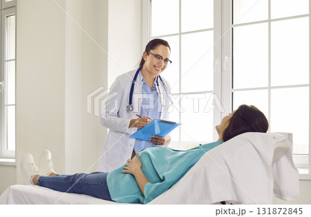 Friendly, smiling doctor with clipboard standing by bed with young pregnant woman 131872845