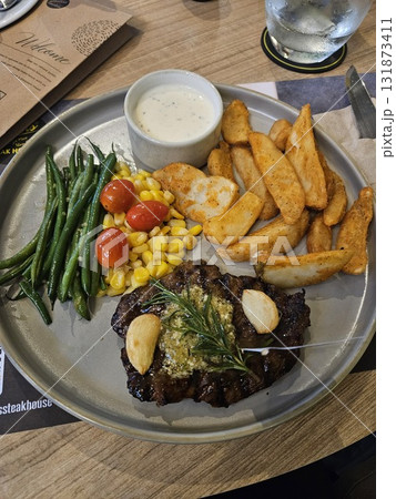 A delicious grilled steak is beautifully plated with roasted garlic, a sprig of rosemary, a side of golden potato wedges, steamed green beans, sweet corn, and bright cherry tomatoes, A delicious grilled steak is beautifully plated with roasted garlic, a sprig of rosemary, a side of golden potato wedges, steamed green beans, sweet corn, and bright cherry tomatoes, 131873411