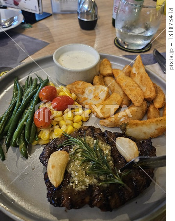 A delicious grilled steak is beautifully plated with roasted garlic, a sprig of rosemary, a side of golden potato wedges, steamed green beans, sweet corn, and bright cherry tomatoes, 131873418