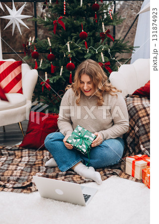 Happy female sitting near Christmas tree preparing gifts for family. A woman looking at a laptop monitor at home during the New Year and Christmas holidays. Full body Happy female sitting near Christmas tree preparing gifts for family. A woman looking at a laptop monitor at home during the New Year and Christmas holidays. Full body 131874923