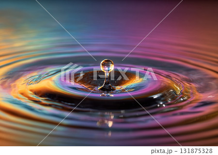 A single water droplet is suspended mid-air above the surface, creating a perfect column and vibrant ripples reflecting warm orange and purple light. 131875328