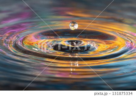 A single water droplet is suspended mid-air above the surface, creating a perfect column and vibrant ripples reflecting warm orange and purple light. A single water droplet is suspended mid-air above the surface, creating a perfect column and vibrant ripples reflecting warm orange and purple light. 131875334