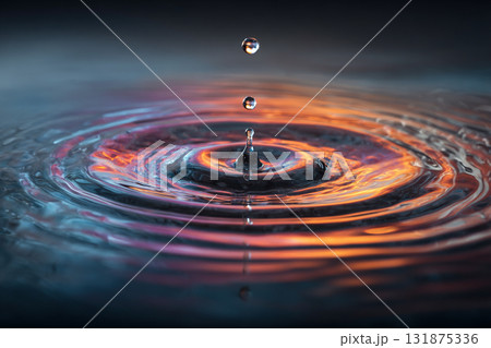 A single water droplet is suspended mid-air above the surface, creating a perfect column and vibrant ripples reflecting warm orange and purple light. A single water droplet is suspended mid-air above the surface, creating a perfect column and vibrant ripples reflecting warm orange and purple light. 131875336