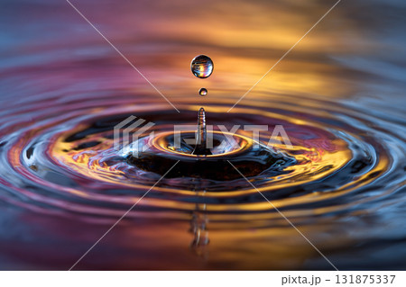 A single water droplet is suspended mid-air above the surface, creating a perfect column and vibrant ripples reflecting warm orange and purple light. 131875337
