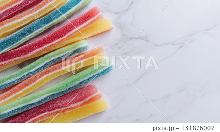 A bright, flat lay of rainbow-striped gummy candy sour belts on a white marble surface, featuring vibrant colors and a sugared texture. 131876007