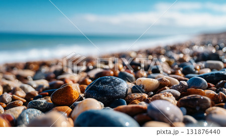 A low-angle, close-up shot of wet, multi-colored pebbles on a beach, stretching toward a blue sea and sky under bright sunlight. 131876440