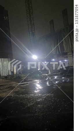 Dimly lit construction site at night with wet reflective ground and bright floodlight creating moody atmosphere 131877054