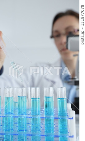 Female scientist in white lab coat pipetting blue liquid into test tubes, microscope visible amid sterile laboratory environment. Medicine, healthcare and science concept 131877078