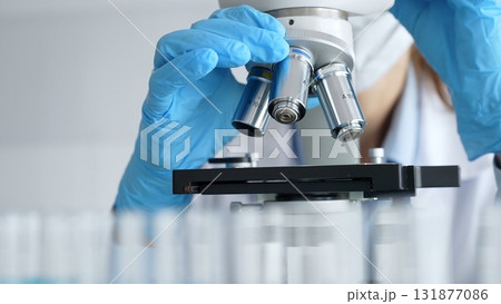 Close up of scientist's gloved hands adjusting microscope, conducting research in laboratory with test tubes containing blue liquid in background. Medicine, healthcare and science concept 131877086