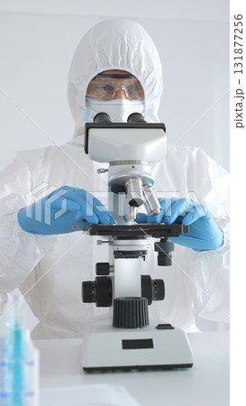 Medical scientist wearing protective suit using microscope while analyzing samples, conducting research in laboratory environment. Close up of test tubes with blue liquid Medical scientist wearing protective suit using microscope while analyzing samples, conducting research in laboratory environment. Close up of test tubes with blue liquid 131877256
